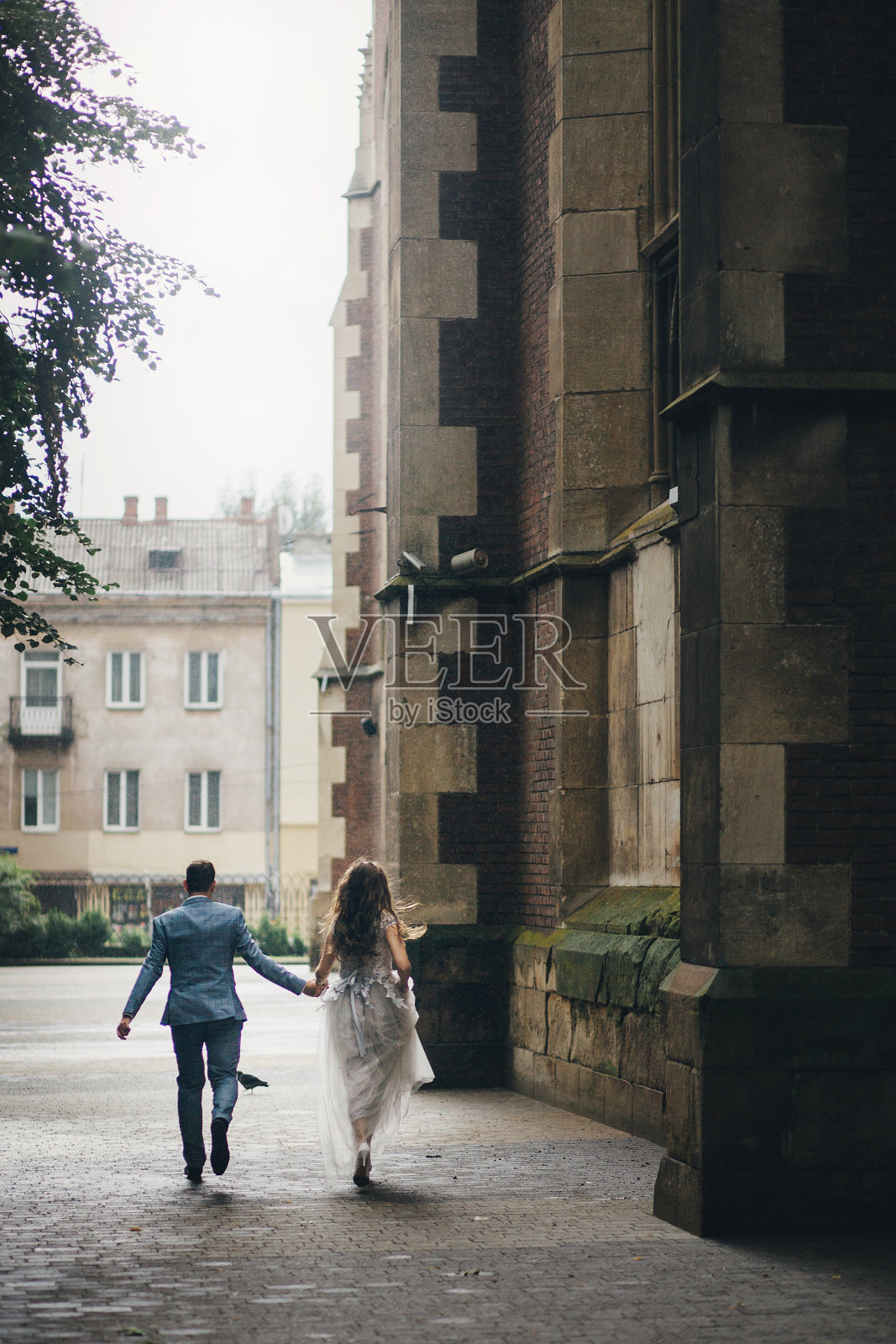 时尚的新娘和新郎在雨街的老教堂背景下奔跑。普罗旺斯的婚礼。美丽的情感婚礼夫妇在雨中在欧洲城市的乐趣。照片摄影图片