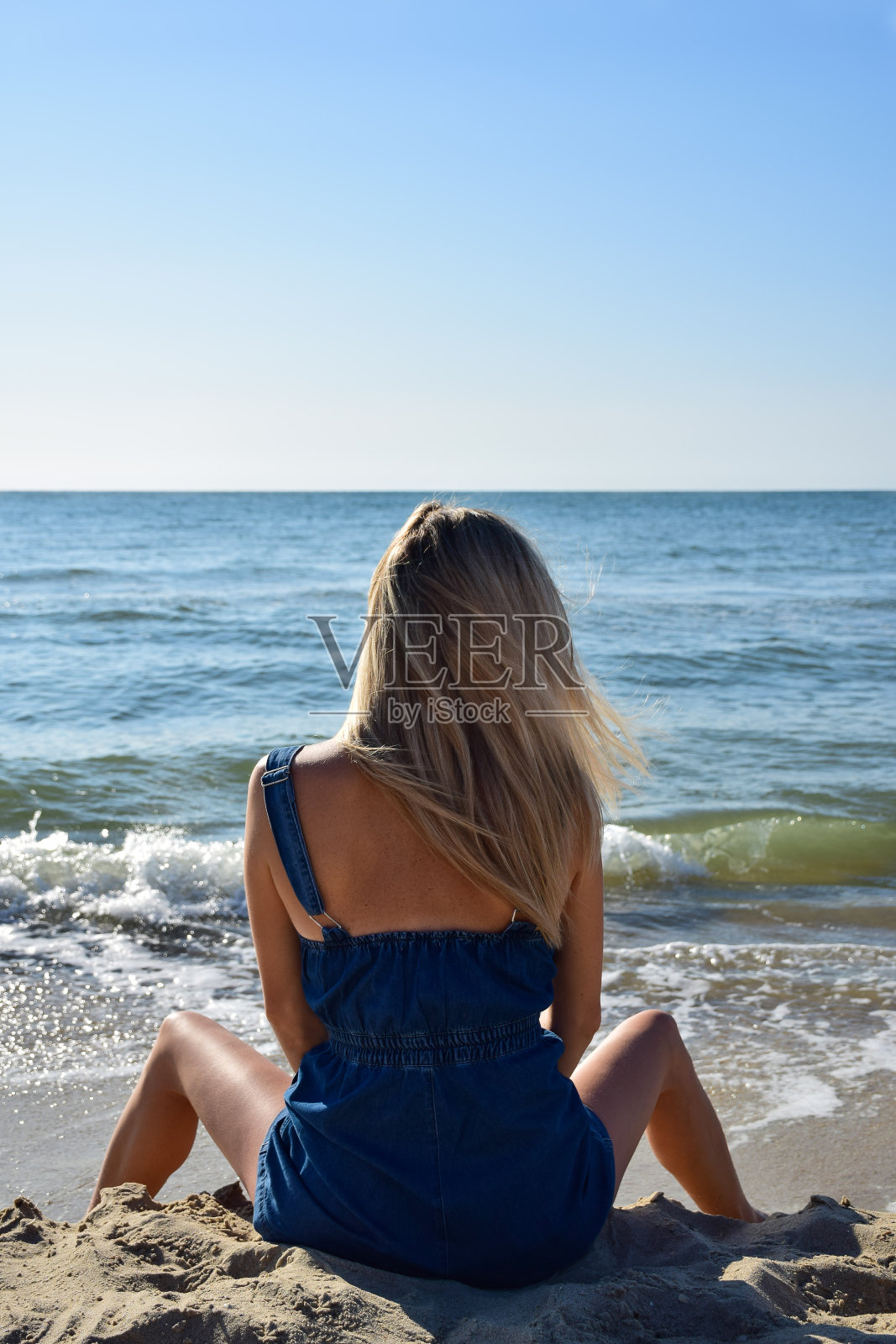 早晨，一个年轻的金发女孩叉开双腿坐在沙滩上。后视图照片摄影图片