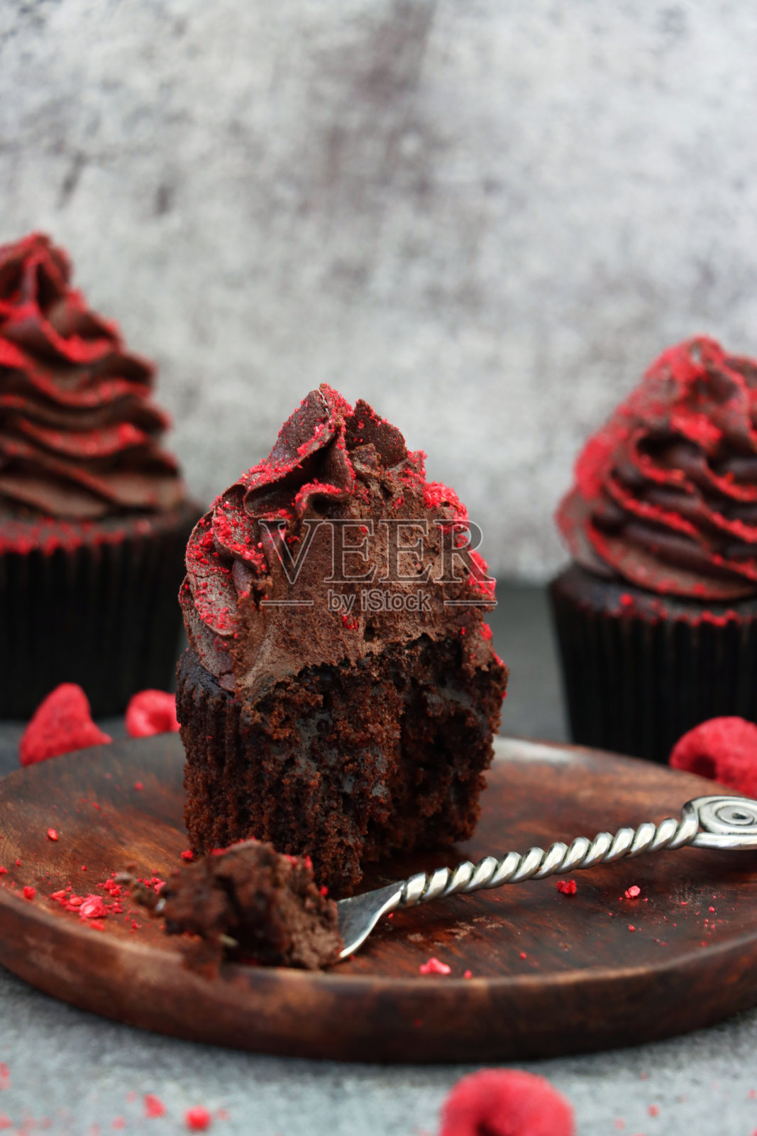 个人自制的巧克力纸杯蛋糕，放在木制的盘子上，用叉子叉住，巧克力黄油糖霜被干燥的覆盆子粉末包裹，没有咬痕，灰色背景，聚焦前景照片摄影图片