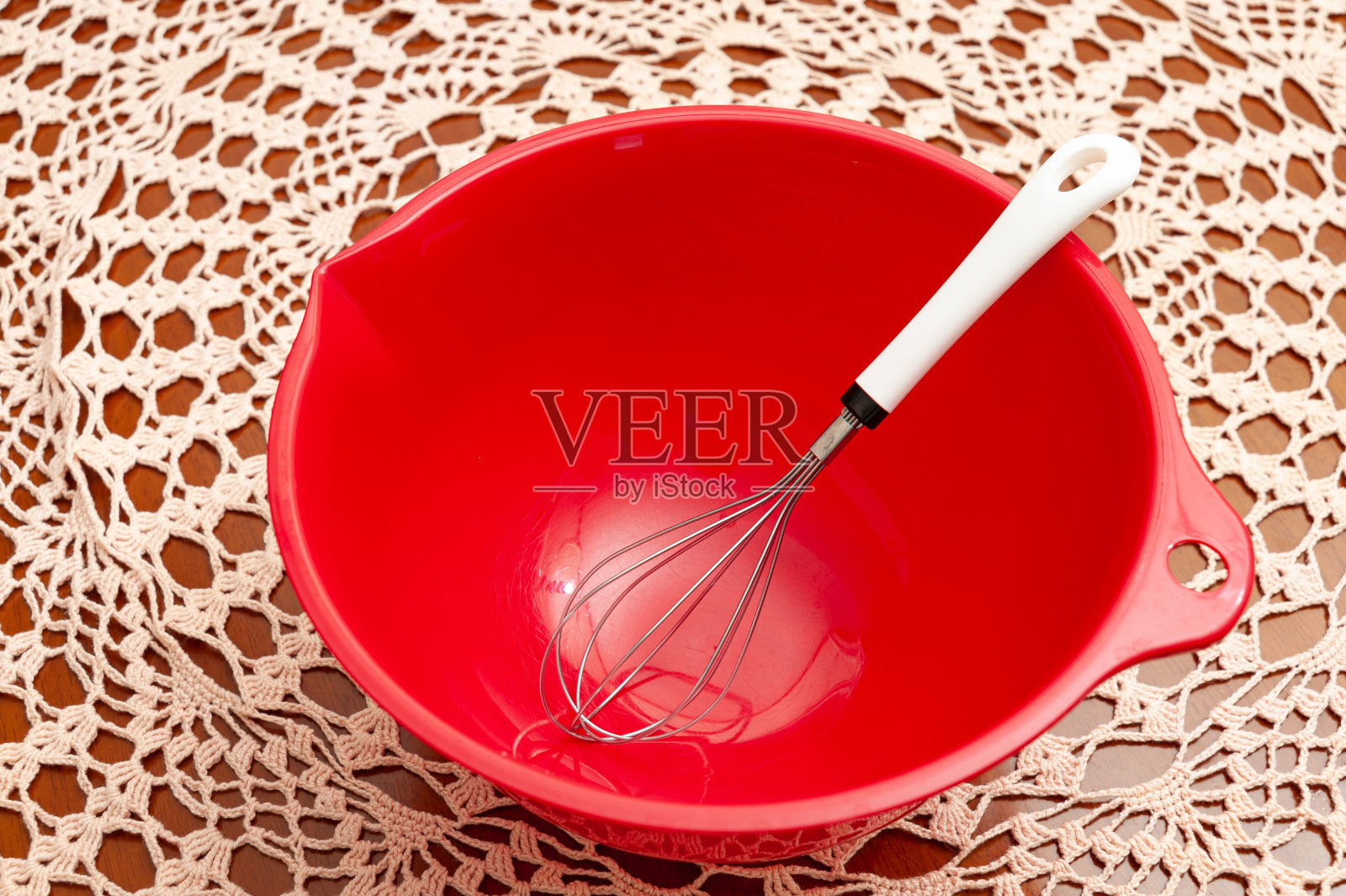 将盛有鸡蛋打蛋器的红色塑料碗倒空。钩针桌布的背景。前视图。特写镜头照片摄影图片