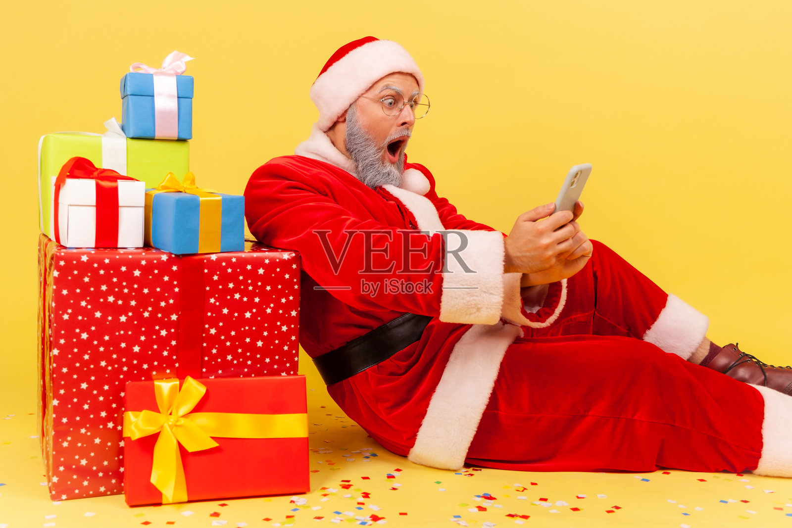 灰色胡子、穿着圣诞老人服装的老人坐在新年礼物旁边，用智能手机读着令人震惊的消息。照片摄影图片