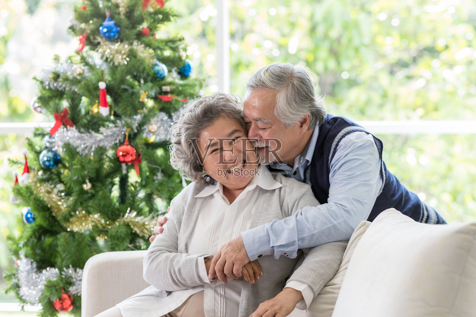 快乐的亚洲老人在圣诞节在家拥抱和亲吻他的妻子。老夫妇坐在沙发上与圣诞树共度时光。圣诞快乐，节日快乐照片摄影图片
