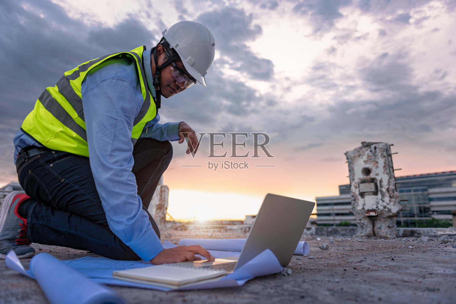 土木工程师穿着高能见度的安全背心和头盔蹲在建筑工地的阳台上看笔记本电脑的图纸细节。翻新概念测量师。照片摄影图片
