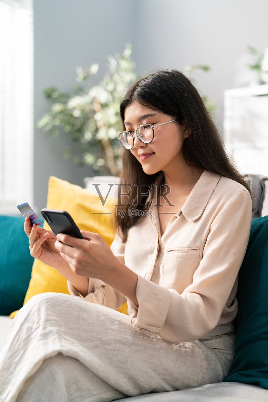 一个亚洲美女购物狂在客厅的沙发上，手里拿着电话和银行卡，信用卡，女人在网上购物，订购新衣服，化妆品，微笑照片摄影图片