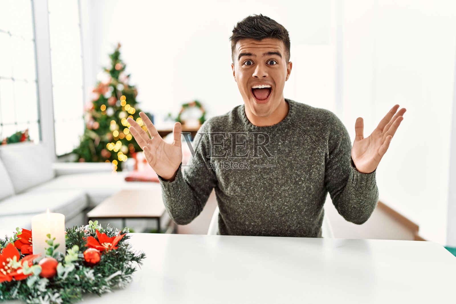 年轻的西班牙裔男子坐在圣诞树旁的桌子上，疯狂地庆祝成功，张开双臂，兴奋地尖叫着。赢家的概念照片摄影图片