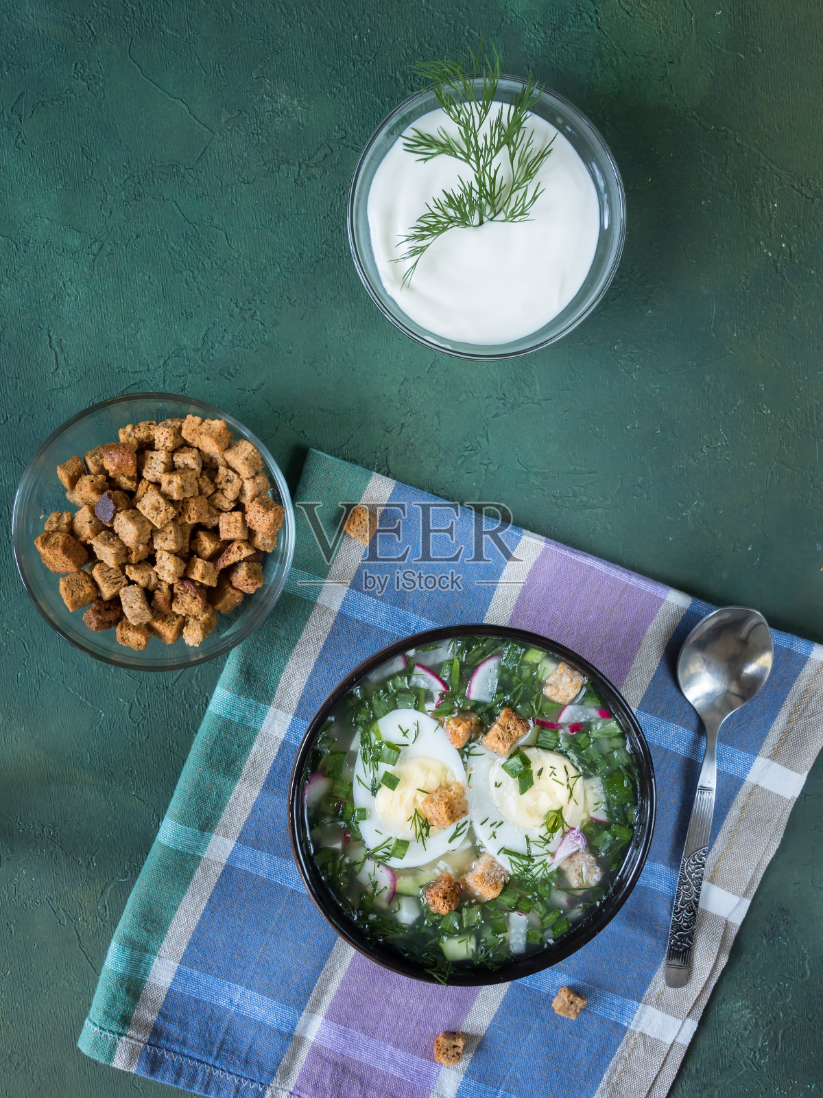 黑色的碗里放着格子餐巾，上面装饰着煮鸡蛋和香草，是传统的寒冷的夏季俄罗斯okroshka汤。桌子上有一盘汤，饼干，一碗酸奶油。绿色背景。平的。垂直照片摄影图片
