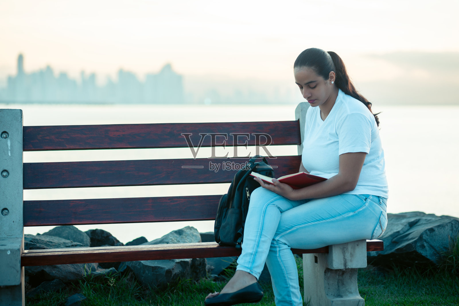 一个女人坐在海边的长椅上读着一本红色的书照片摄影图片