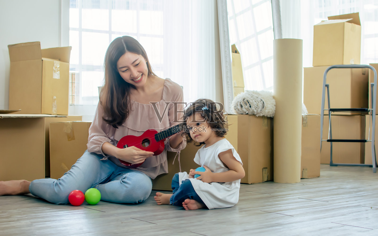 美丽的成年亚洲母亲和可爱的小女儿微笑着，玩着尤克里里，一起唱歌，幸福和乐趣，坐在新房子或公寓的地板上，打开棉花盒。照片摄影图片