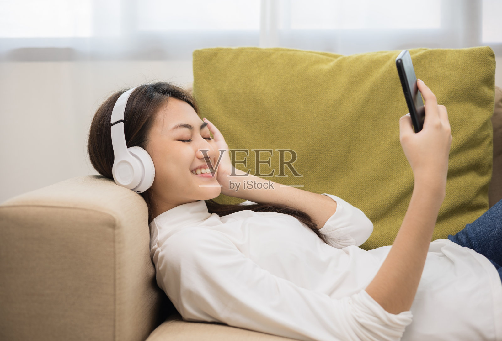 在沙发上放松。美丽年轻的亚洲女人躺在沙发上，用耳机听着音乐。她有节奏的生活方式在客厅在家里早上。照片摄影图片