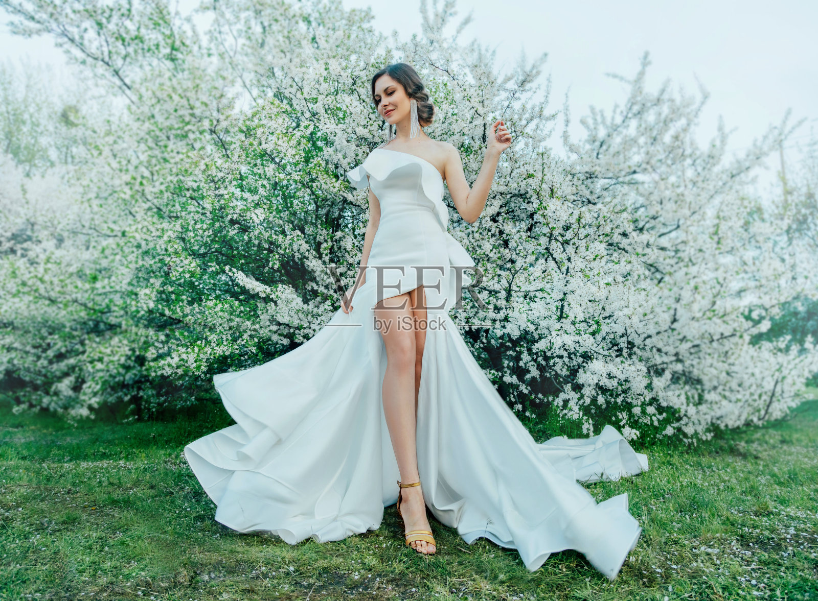 幻想中的女子穿着白色优雅时尚的长裙，漫步在春绿盛开的樱花园里。快乐开朗的女孩公主新娘裙布飞扬，随风飘动。性感的裸腿照片摄影图片