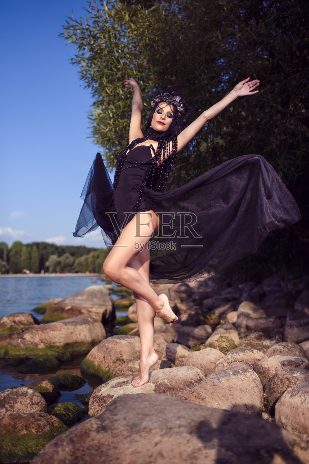 跳舞的想法。年轻的黑发女芭蕾舞演员在岩石海岸上穿着黑色连衣裙，戴着贝壳装饰的王冠和珍珠项链。垂直的图像照片摄影图片