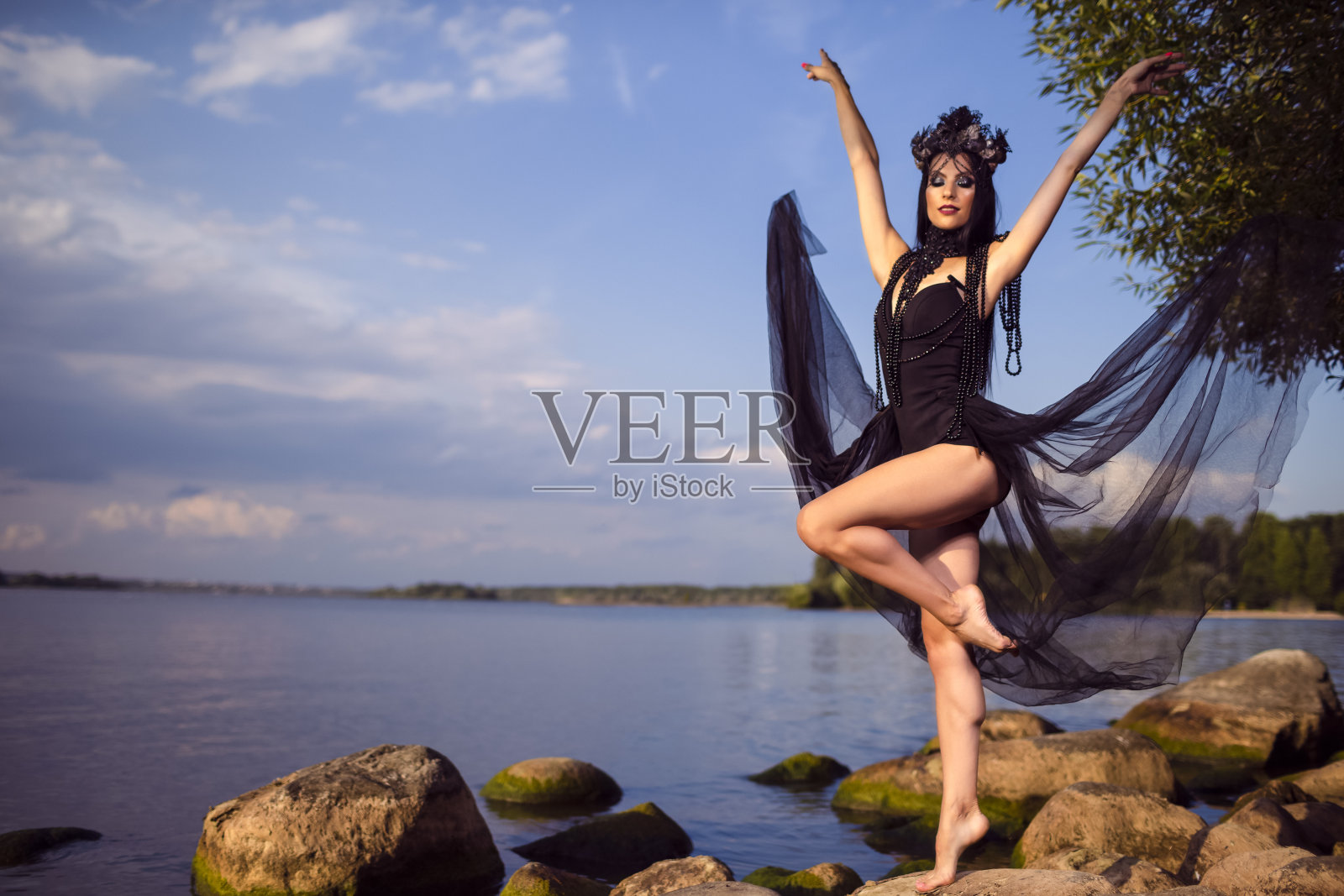 概念的舞蹈。身穿黑色连衣裙的年轻黑发女芭蕾舞演员在岩石海岸上跳舞，戴着贝壳装饰的王冠和珍珠项链。水平的作文照片摄影图片