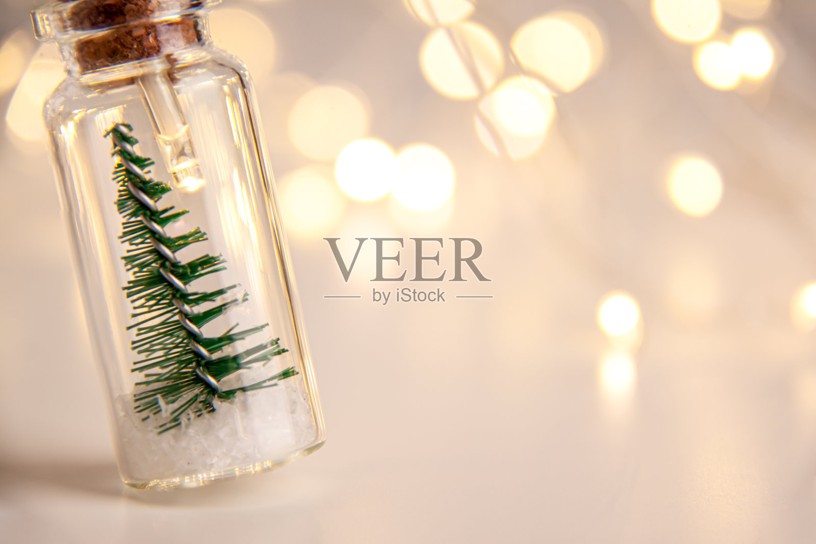 靠近发光的灯装饰的玻璃瓶与软木塞，里面有小的绿色圣诞树的人造雪。假期新年概念，左边有复印空间和散景照片摄影图片