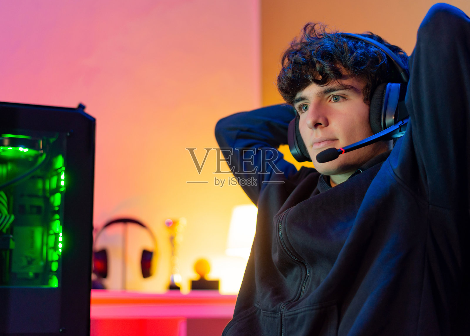 年轻的白人青少年玩家带着耳机靠在椅背上，手臂在脑后，满意的脸。照片摄影图片