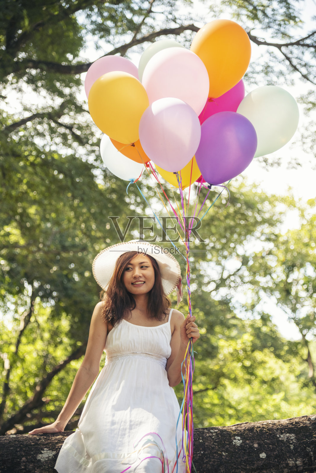 欢快的美女拿着气球坐在绿色公园的大树下，心情舒畅。女人手握充满活力的气球在生日派对上玩快乐的时间夏天在户外的阳光照片摄影图片