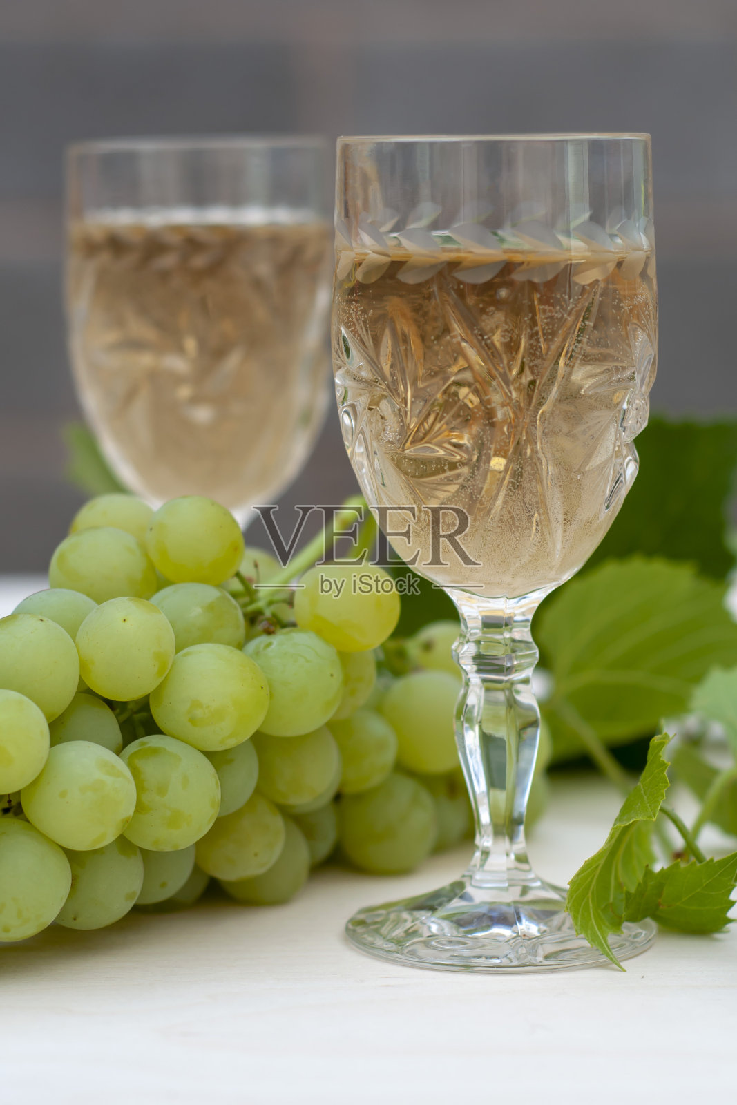 两杯白葡萄酒，几串葡萄和一根年轻的藤蔓放在浅色的木制背景上。一个垂直的快照。照片摄影图片