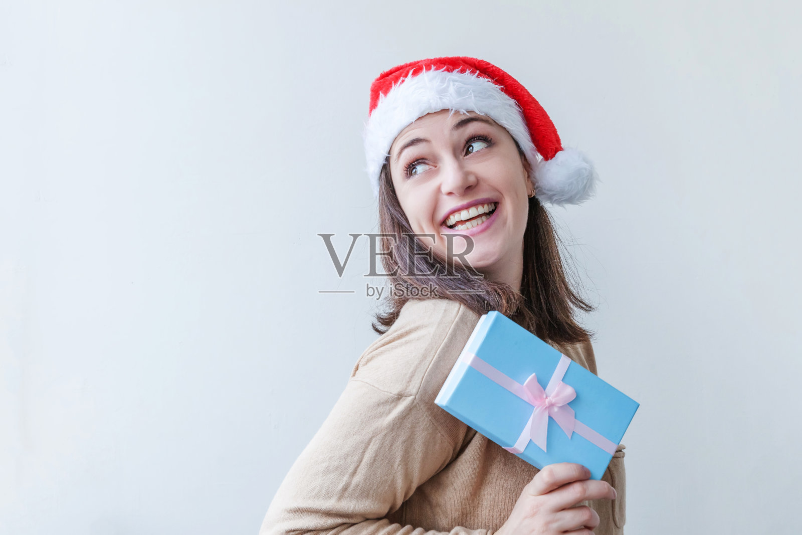 美丽的女孩戴着红色的圣诞老人的帽子，手里拿着蓝色的礼盒孤立在白色的背景上。年轻女人的肖像，真实的情感。圣诞和新年假期概念快乐照片摄影图片
