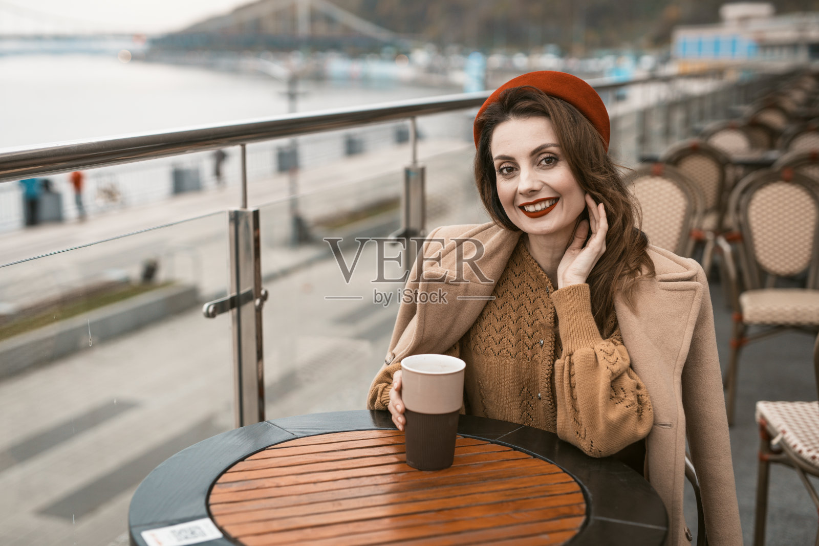 快乐的年轻女孩坐在户外街道餐馆或阳台看着微笑着拿着咖啡杯。穿着秋日大衣和红色贝雷帽的时尚年轻女人的肖像在户外照片摄影图片