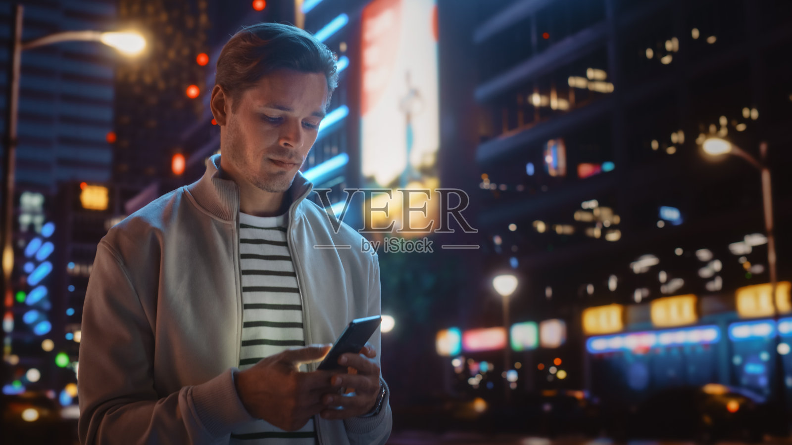 一名男子在霓虹灯照亮的夜城街道上使用智能手机。微笑的时尚男人使用手机，社交媒体，网上购物，在约会App上发短信。照片摄影图片