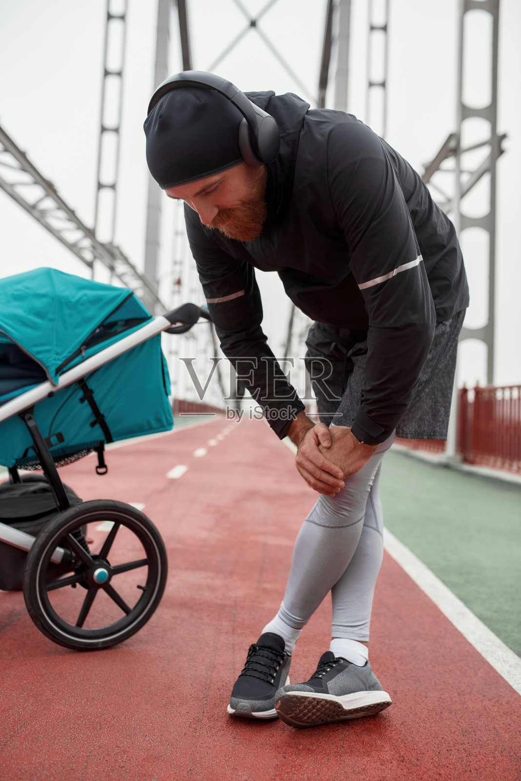 疲倦的运动年轻父亲戴着耳机在户外训练后休息与婴儿车在一个阴天在城市照片摄影图片