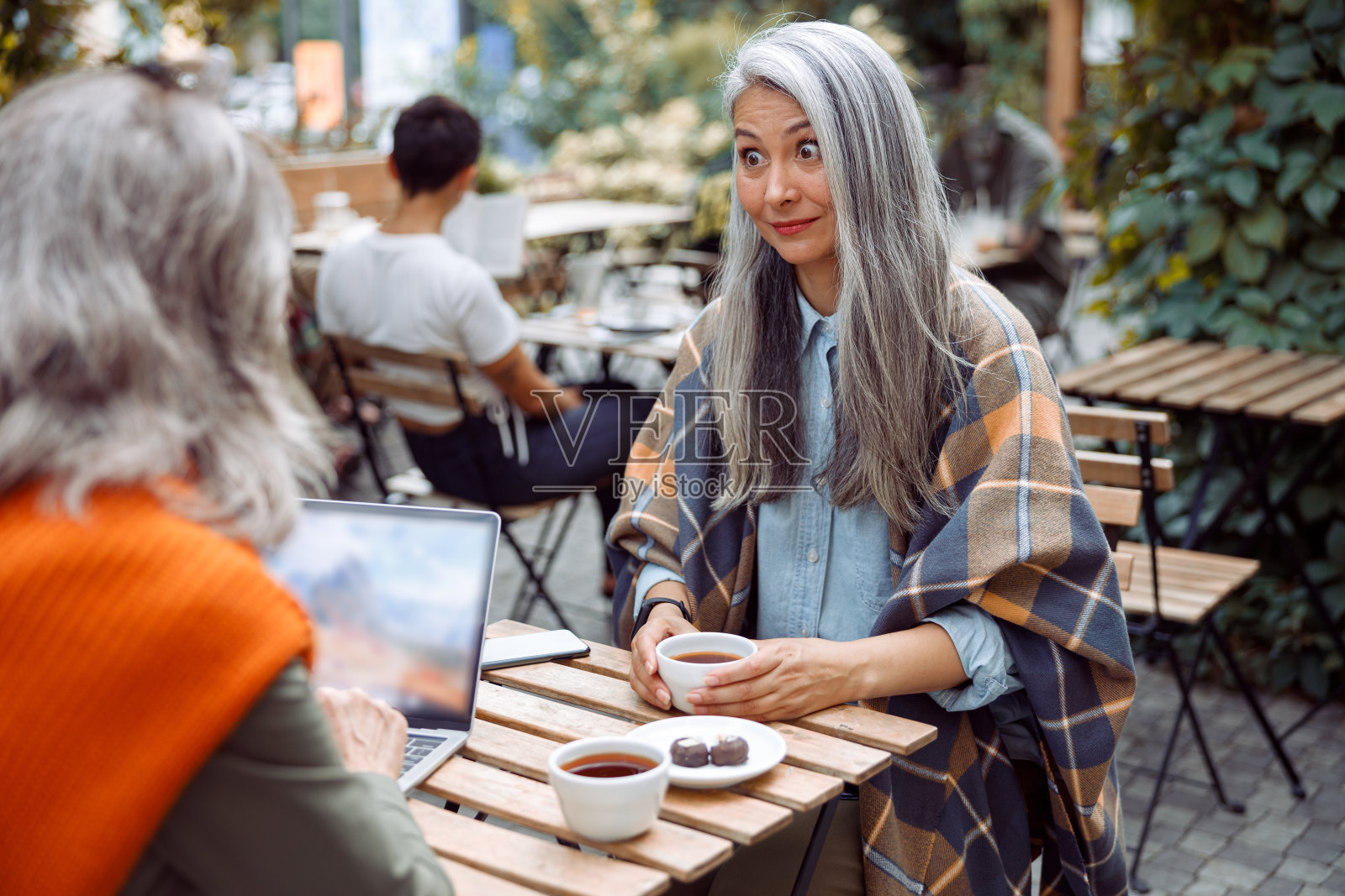 在街边咖啡馆的小桌子上，一位成熟的亚洲女士惊讶地看着正在使用笔记本电脑的朋友照片摄影图片