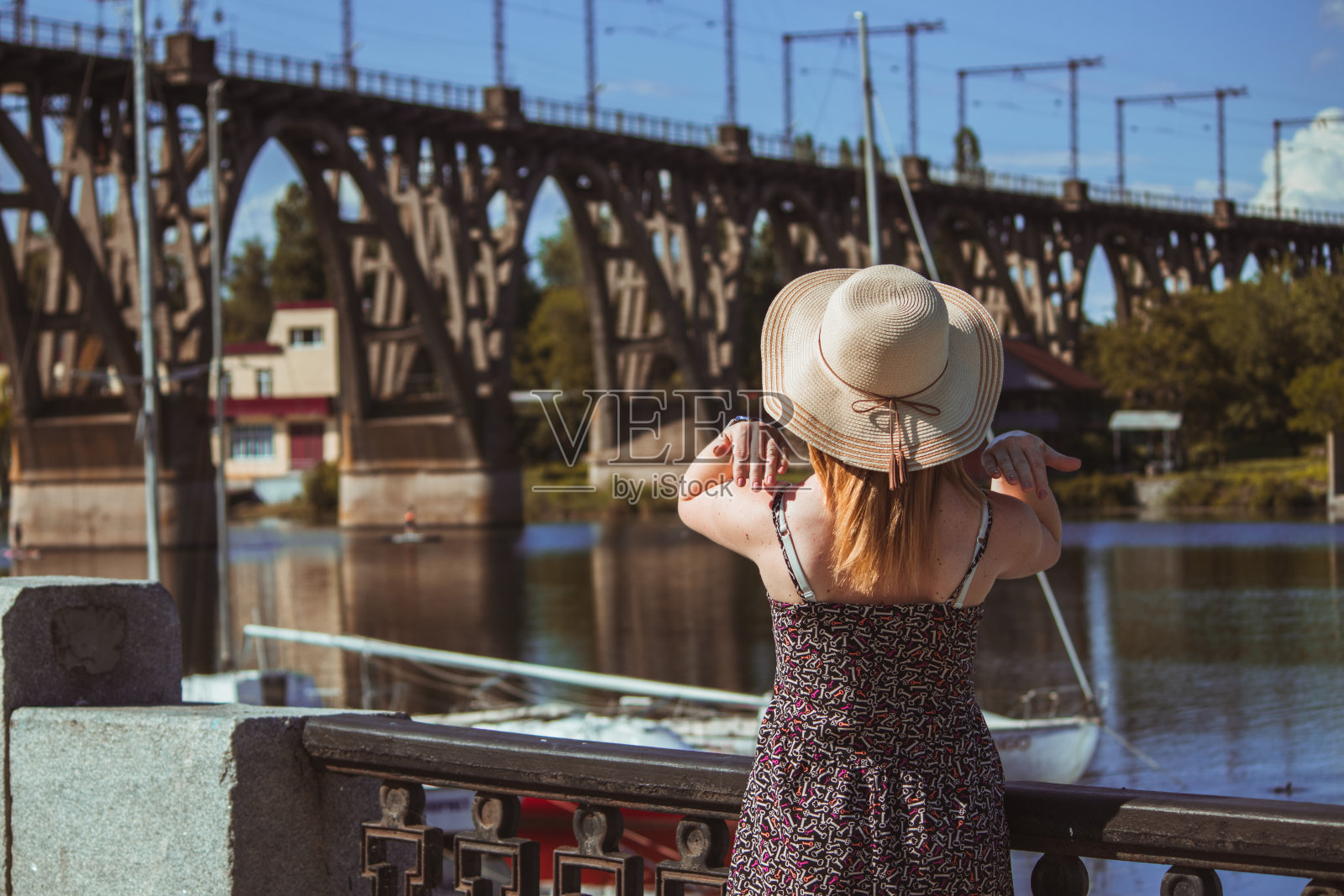 一个身穿连衣裙、头戴夏帽的女孩站在河堤上，把头发拉直。一个阳光明媚的夏日，一个女孩站在游艇俱乐部和铁路桥旁边。照片摄影图片