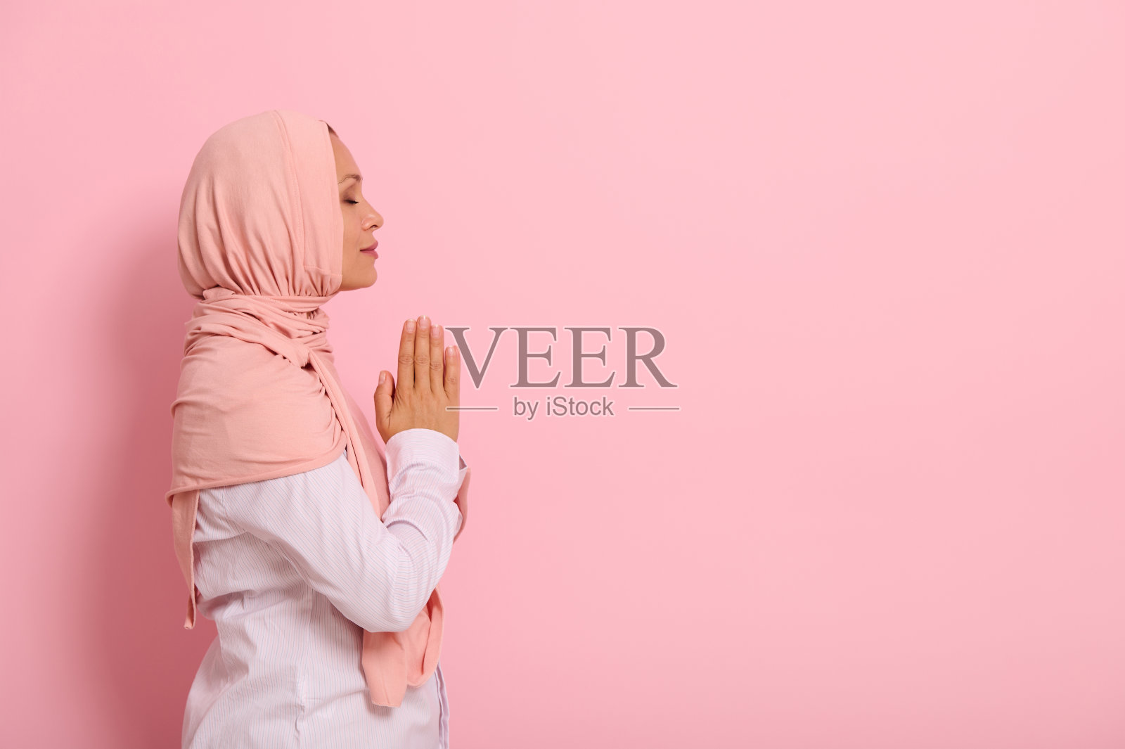 穆斯林阿拉伯妇女身穿粉红色头巾和严谨的服装，双手合十于胸前祈祷的侧面肖像，在彩色背景上，有文字空间照片摄影图片
