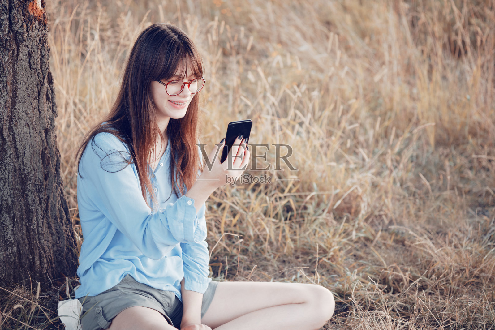 一个戴着眼镜的少女坐在草坪上的一棵树旁看着电话。照片摄影图片