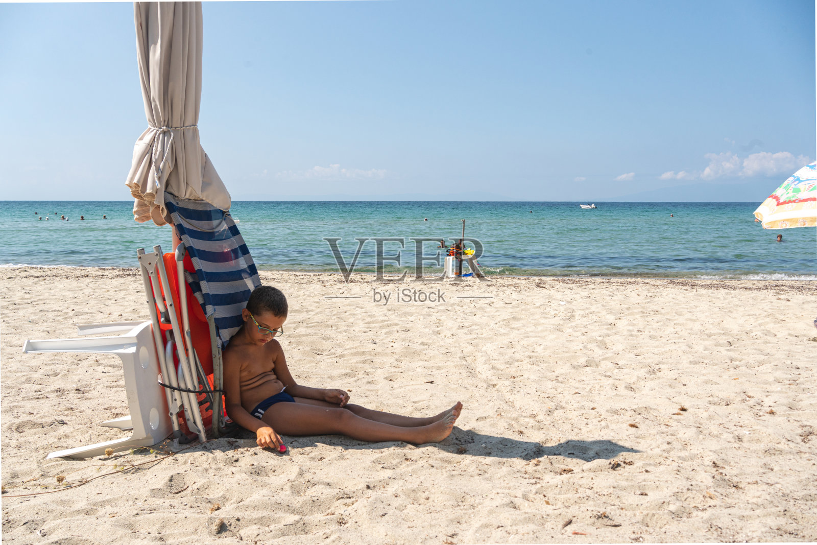 一边等着朋友们一起在沙滩上玩耍，男孩坐在阳伞的阴影下照片摄影图片