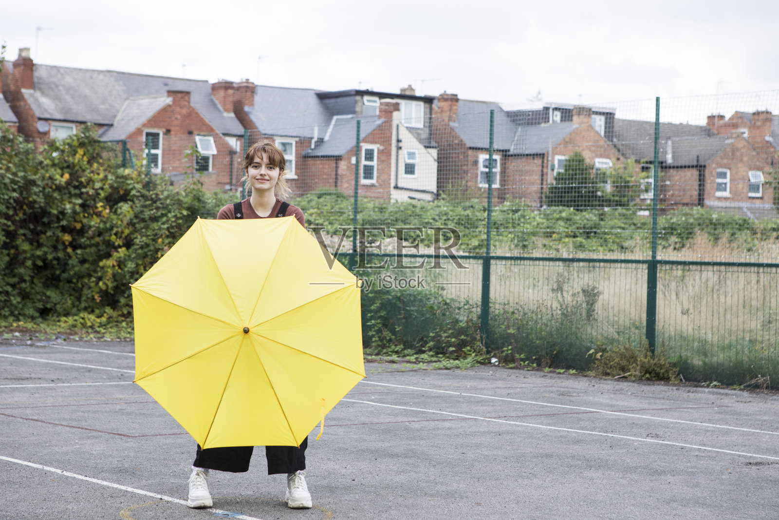 在一个废弃的网球场上，一名年轻女子拿着一把黄色的雨伞，微笑着看着镜头照片摄影图片