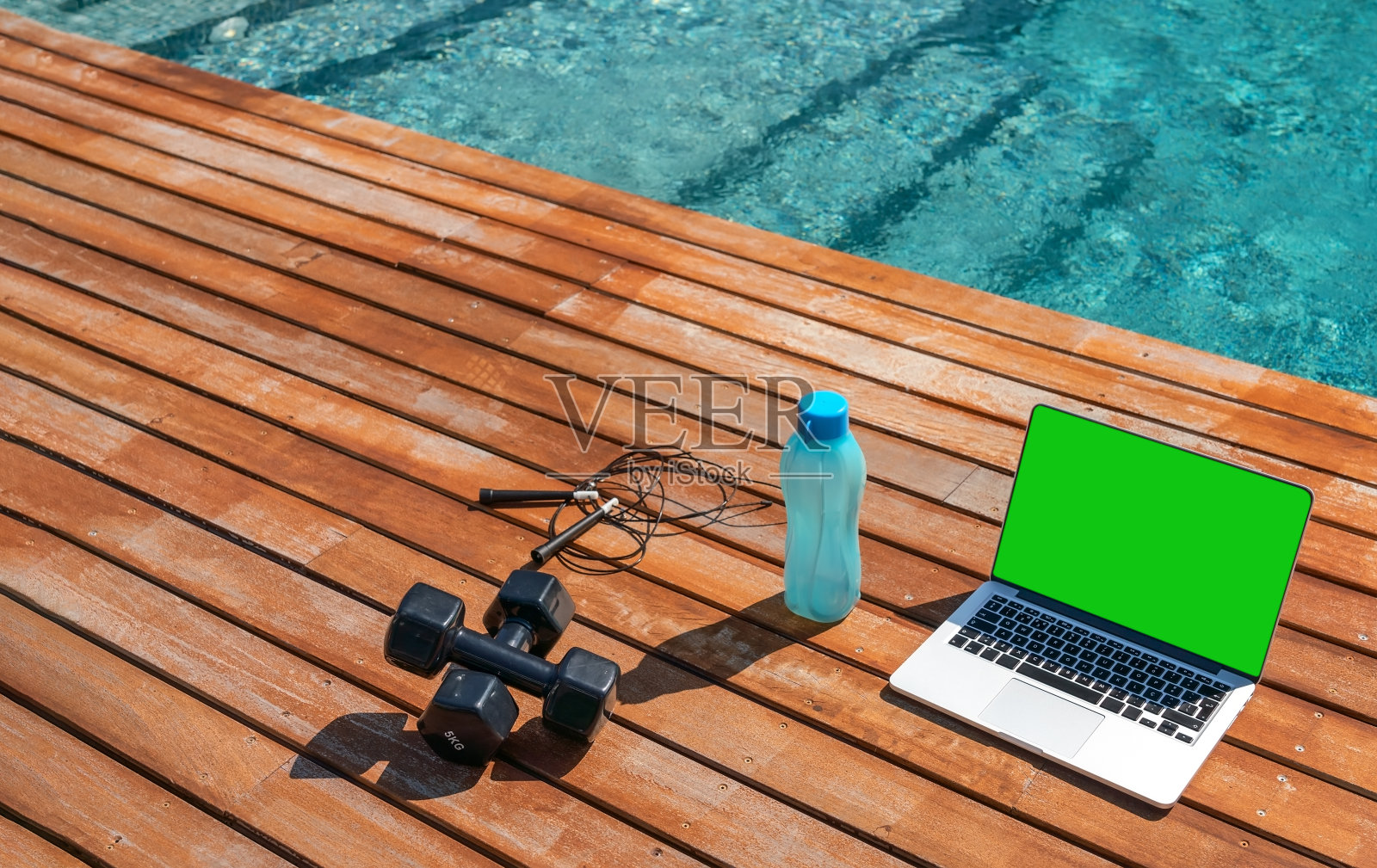 一个哑铃，一根跳绳，一瓶水和一台用于在游泳池附近在线锻炼的电脑的健身概念图。近,外面。照片摄影图片