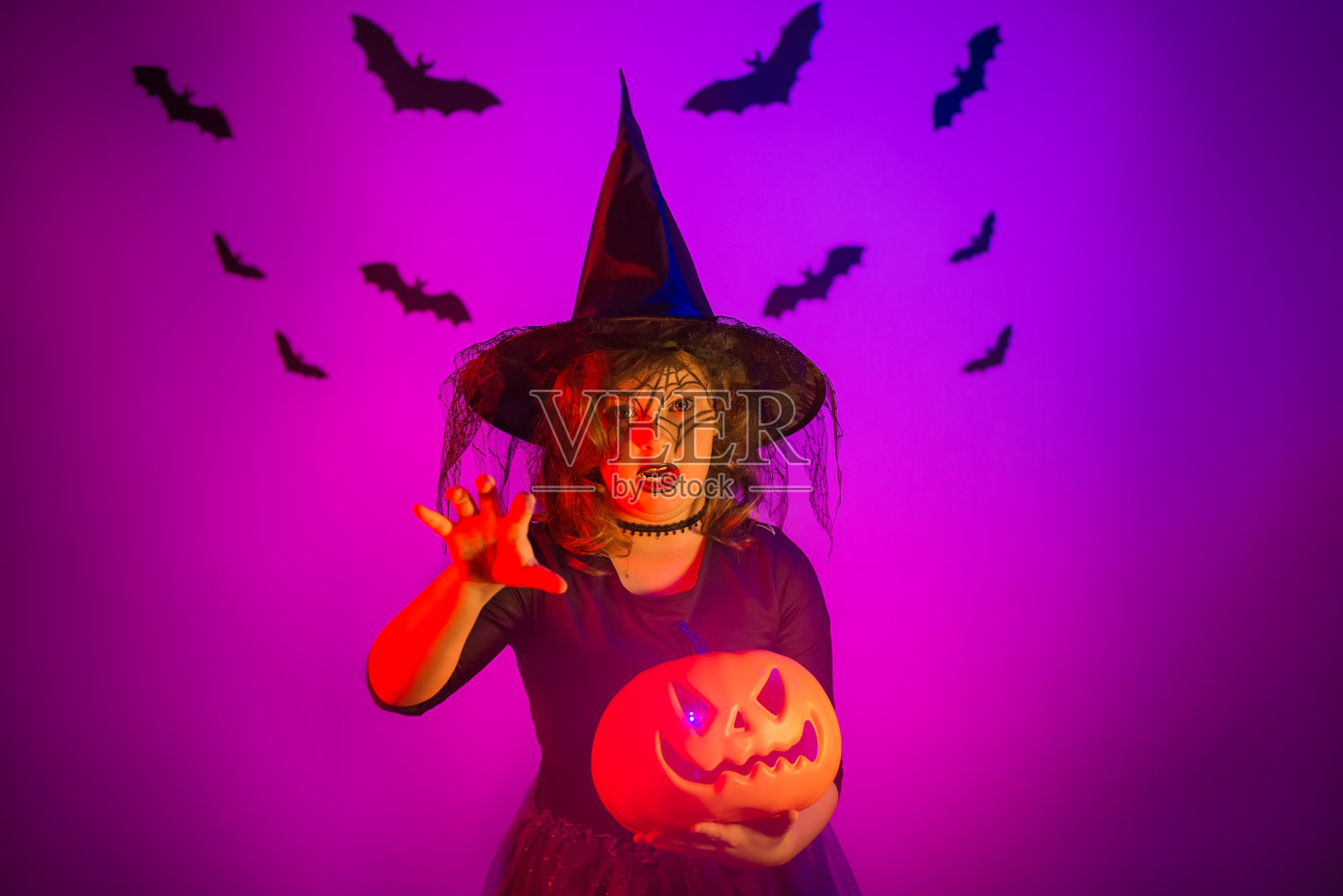 一个十几岁的女孩穿着万圣节女巫服装，脸上画着南瓜和蜘蛛网。工作室在美丽的霓虹灯紫色照明。万圣节快乐概念照片摄影图片