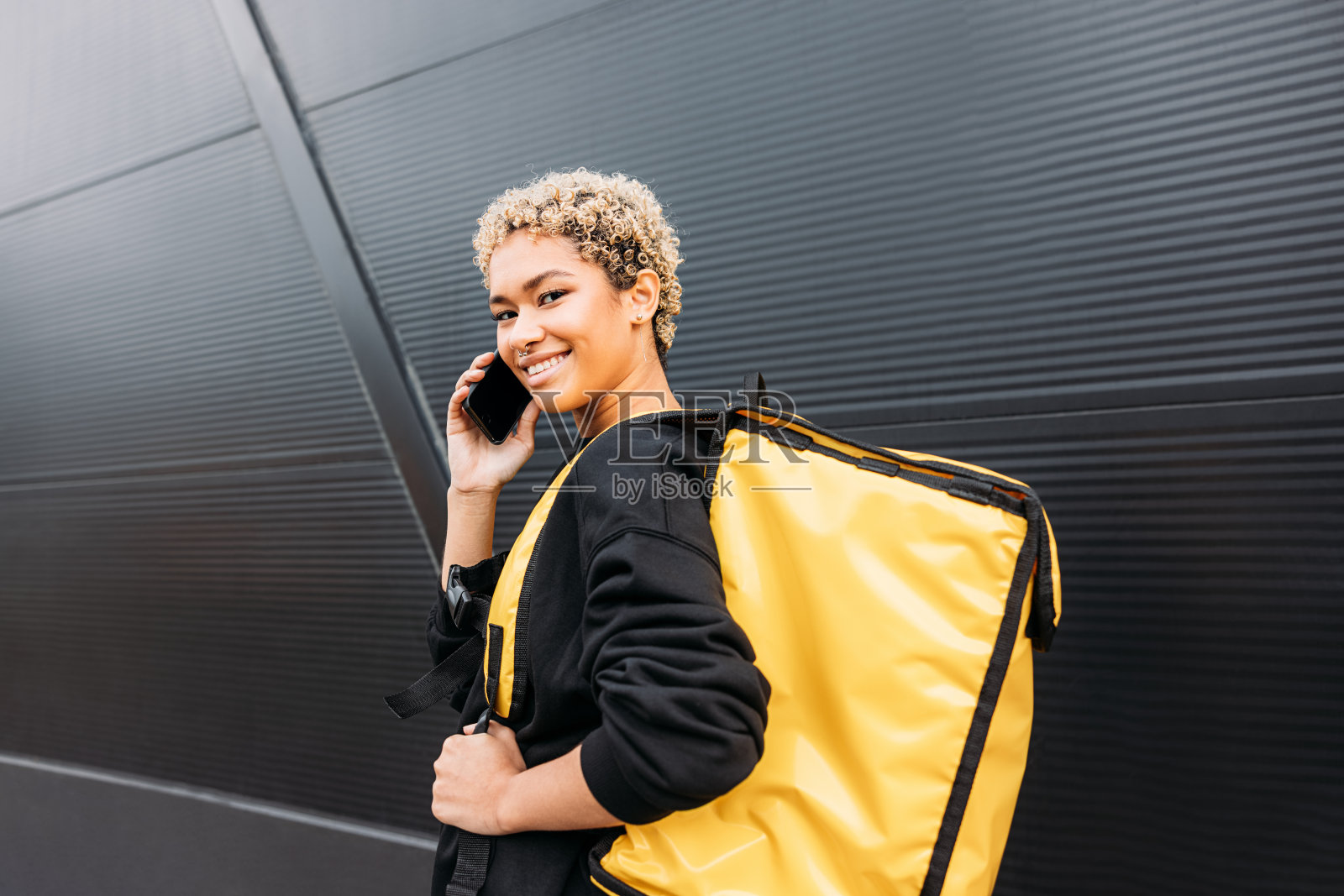 背着背包的女快递员一边用手机聊天，一边看着黑色墙壁上的摄像头照片摄影图片
