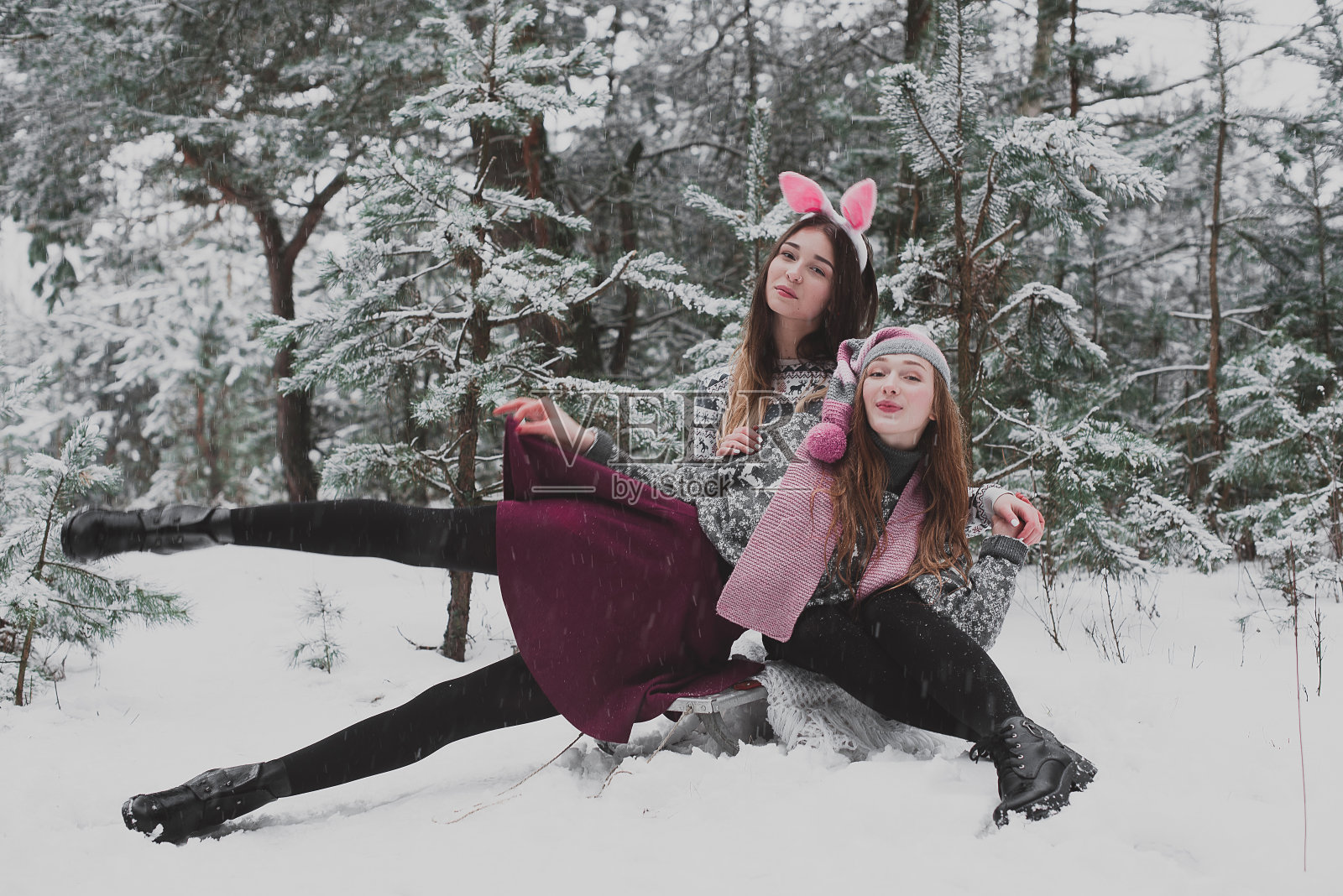 两个年轻的潮女朋友在一起。近距离拍摄两姐妹的时尚肖像，她们戴着粉红色的帽子，戴着兔子耳朵和毛衣，在雪地里，最好的朋友情侣在户外玩耍照片摄影图片