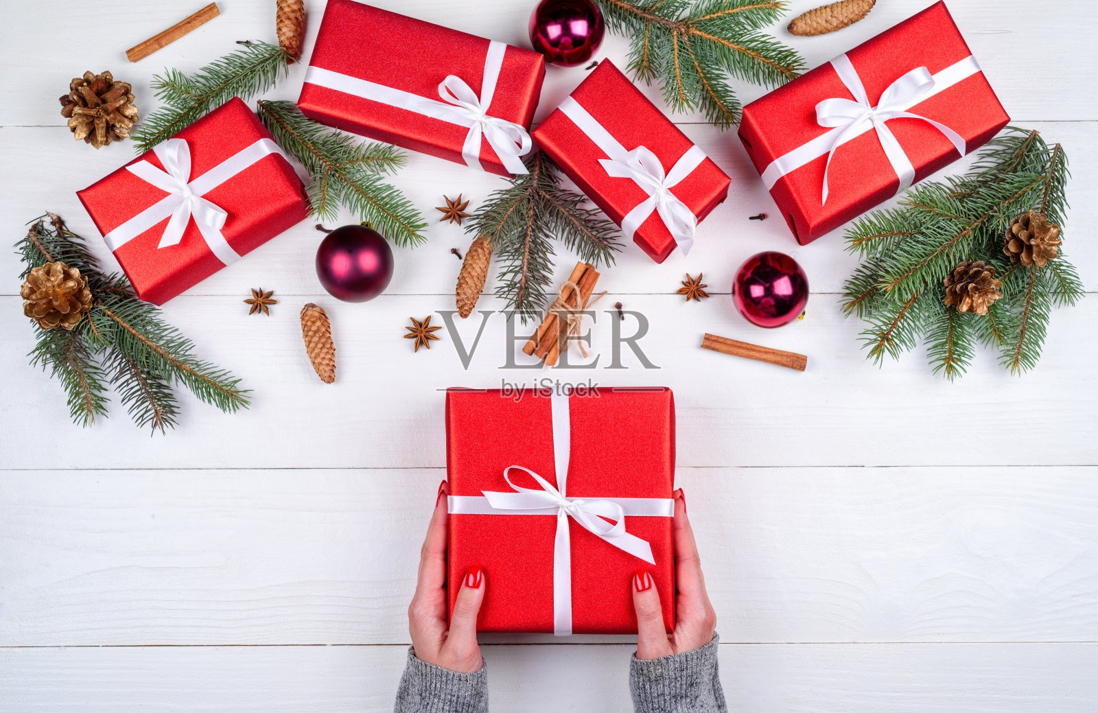 手持红色圣诞礼盒白色木制背景与圣诞装饰(礼物，冷杉树，松果，肉桂棒和八角)，复制空间。平铺，俯视图照片摄影图片