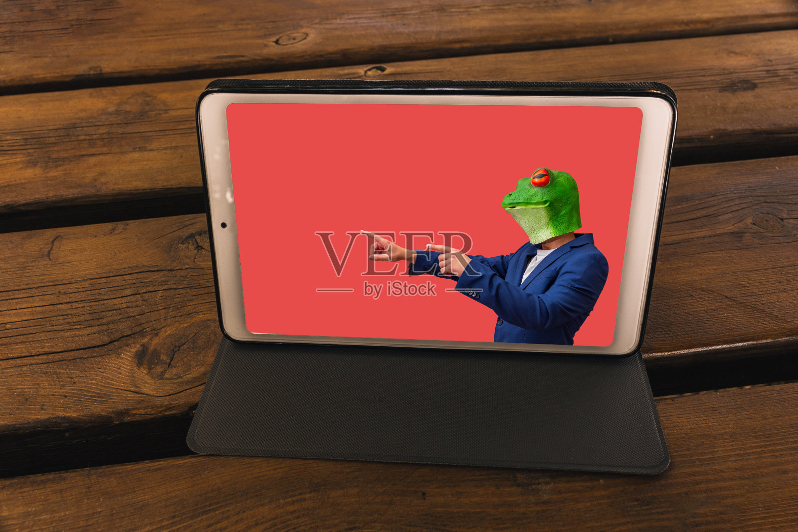 在花园的木桌上的一块平板上，红色背景上有一个戴着青蛙面具的男人照片摄影图片