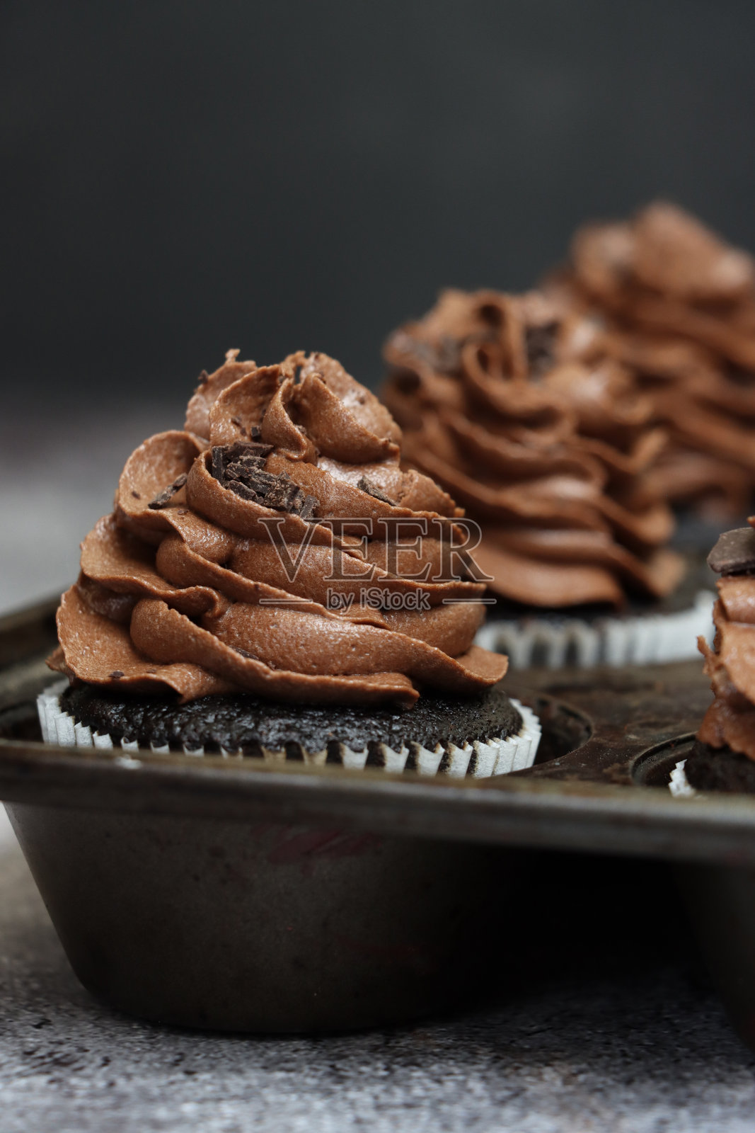 图片中的马芬蛋糕罐，里面装着自制的，纸制蛋糕盒中的巧克力纸杯蛋糕，巧克力黄油糖霜，上面有巧克力块，灰色背景，前景聚焦，复制空间照片摄影图片