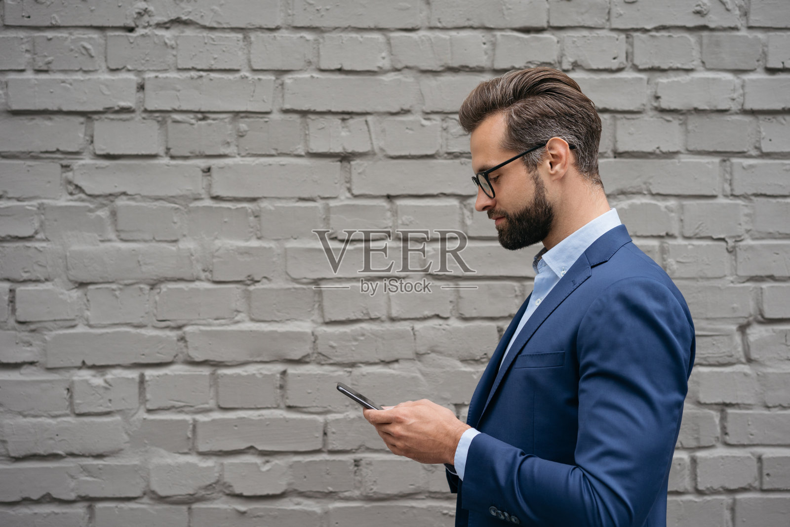 商人穿着西装，戴着时尚的眼镜，拿着手机，交流线上孤立在灰色背景上。大胡子男子在用手机应用，在网上购物，在户外散步照片摄影图片