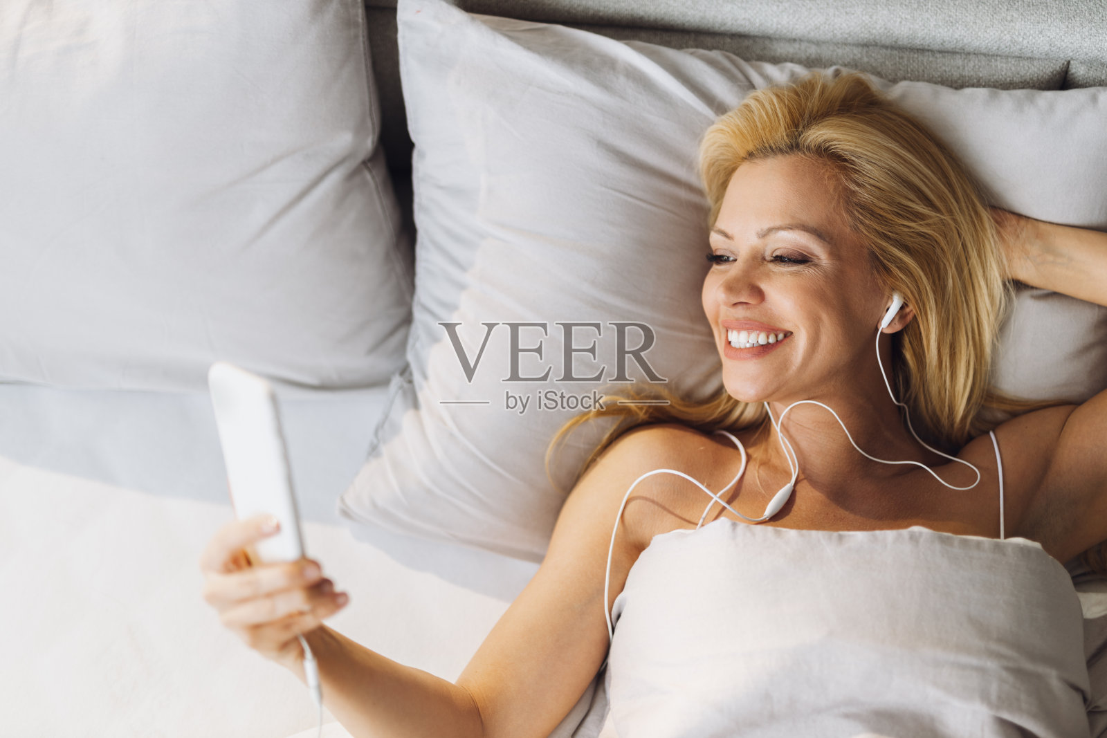 上图为快乐的女人在床上用智能手机听音乐照片摄影图片