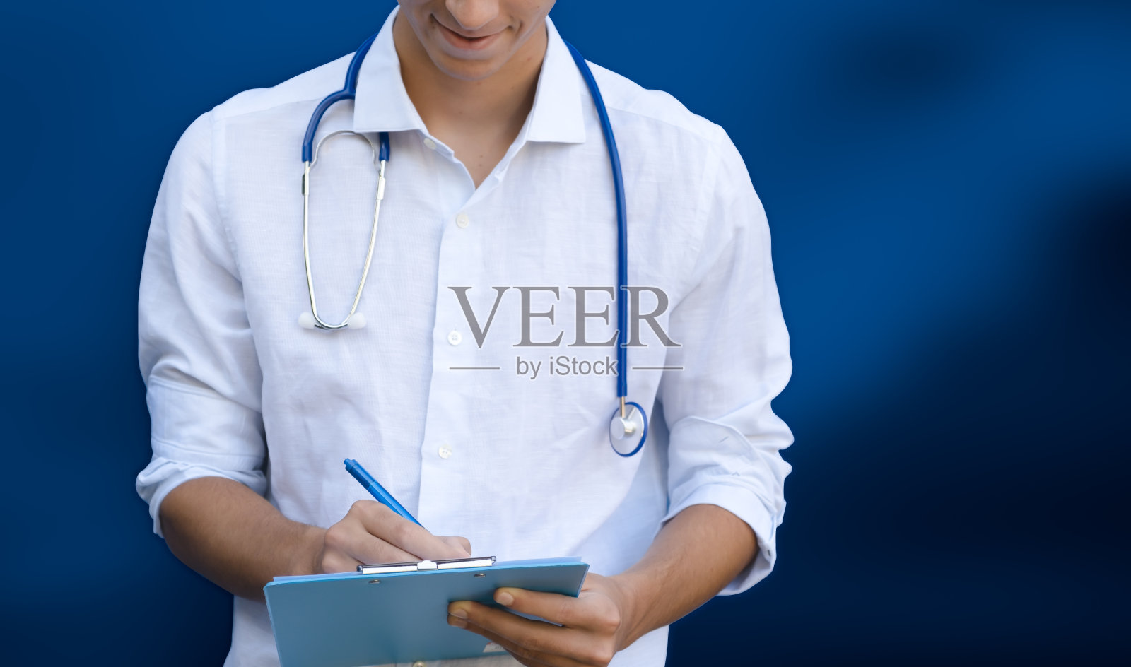 年轻的医生在写字板上写字。蓝色背景与复制空间照片摄影图片