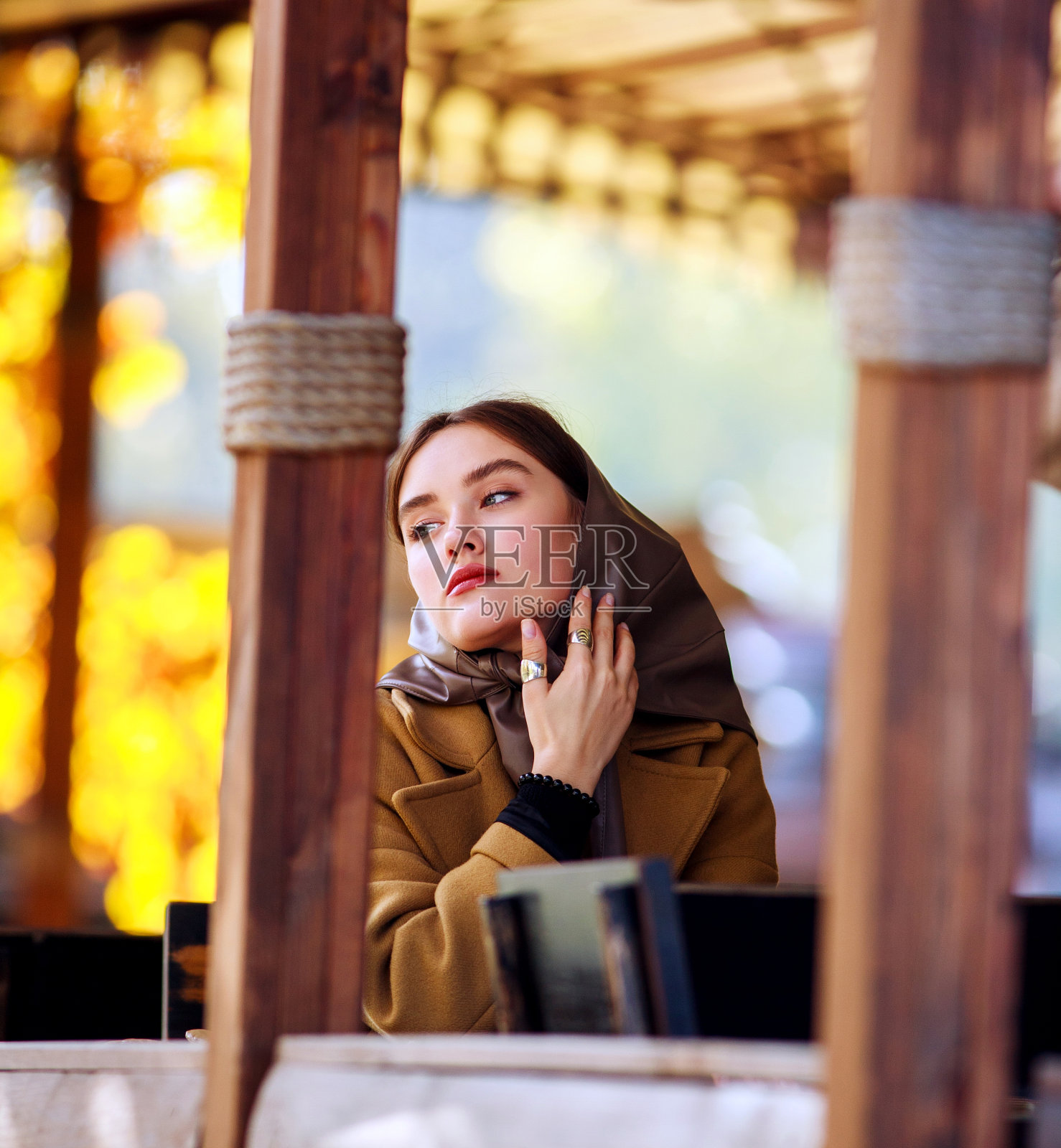 秋天，在木制操场上的咖啡馆里，穿着米色外套、戴着头巾的复古时尚的年轻女子照片摄影图片