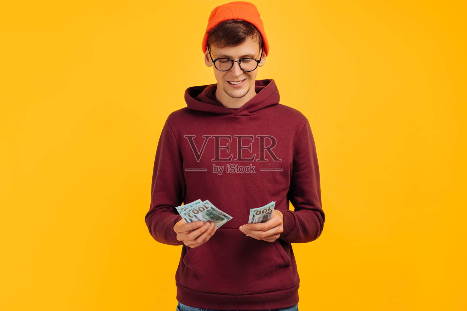 英俊的家伙戴着一顶橙色的帽子，戴着一副眼镜，穿着一件红色的运动衫，手里拿着一把美元的扇子照片摄影图片