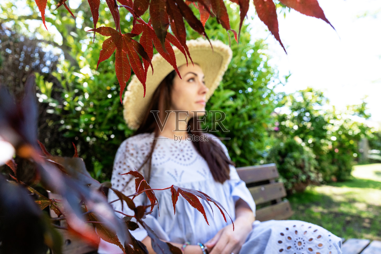 迷人沉思的年轻怀孕黑发，时尚的淡紫色裙子和草帽坐在长椅旁边的树枝在夏季花园照片摄影图片