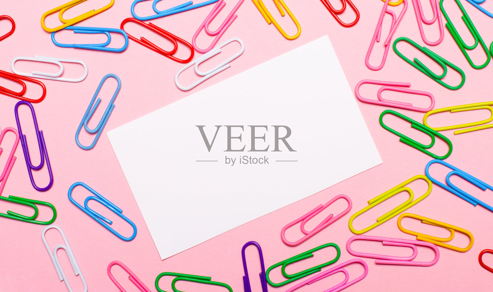在浅粉色的背景上，色彩鲜艳的回形针和一个白色的空白卡片，在中心插入文本。模板。本空间照片摄影图片