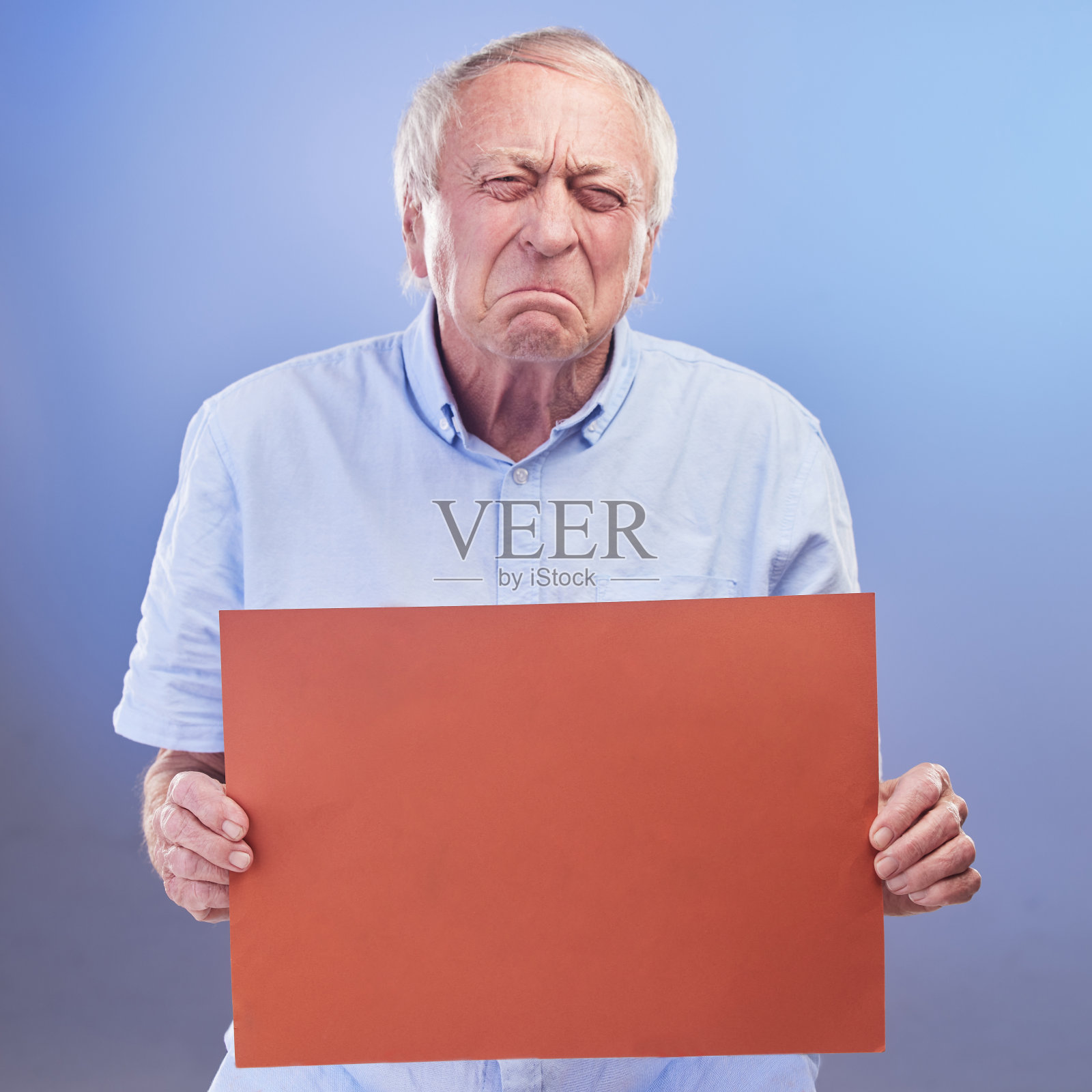 摄影棚拍摄的是一位年长的男子拿着一块空白的牌子，在蓝色的背景下看起来很不开心照片摄影图片