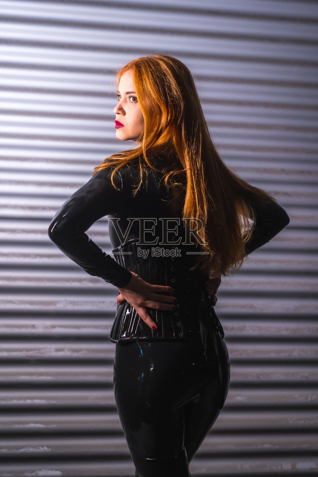 红头发的女孩穿着乳胶，黑色上衣和紧身裤，在金属背景下，在她的背上摆姿势照片摄影图片