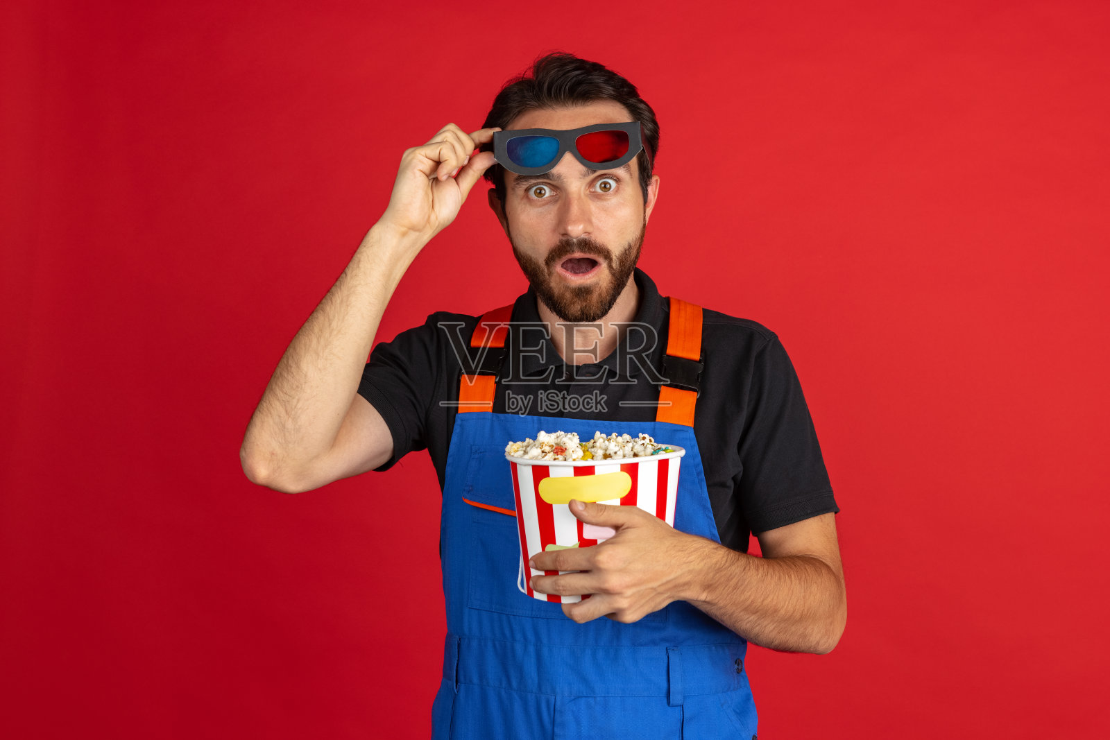 惊讶的胡子年轻男子，男性汽车修理工或钳工穿着蓝色工装裤孤立在红色的工作室背景。面部情绪的概念照片摄影图片