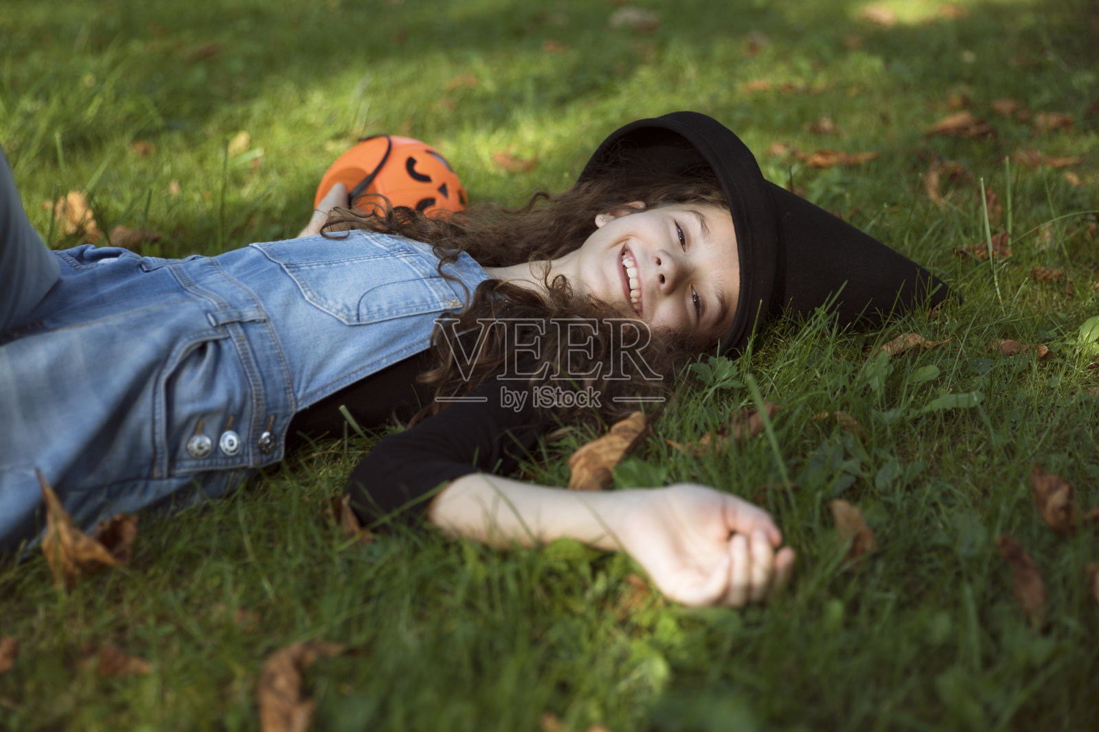 快乐可爱的小女孩戴着女巫长长的黑帽子，躺在草地上，手里拿着一个橙色的大锅。照片摄影图片