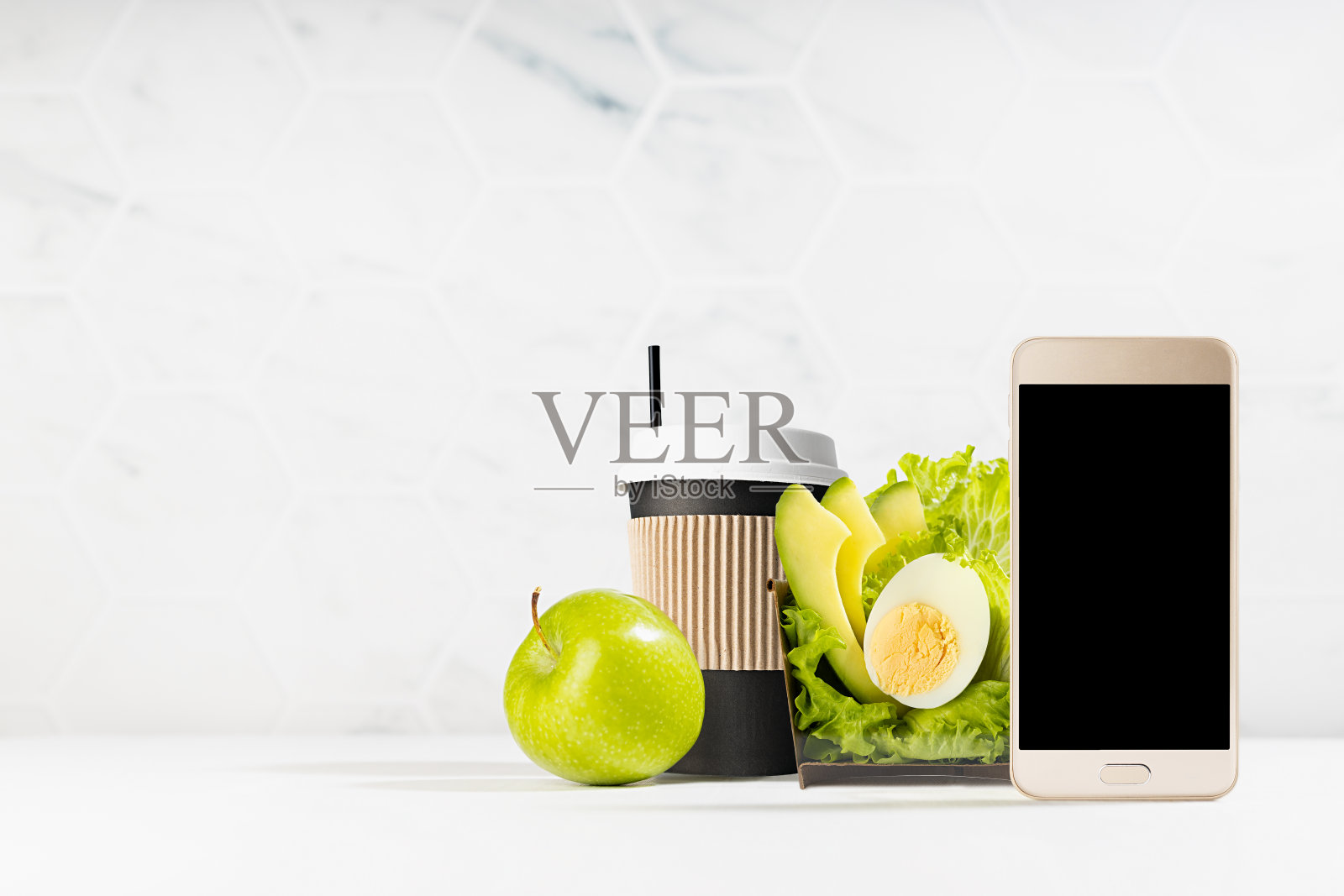 健康早餐外卖-一套绿色沙拉，一杯咖啡，绿色苹果在白色的内部与空白屏幕手机。为外卖服务或外卖餐厅做广告的模型。照片摄影图片