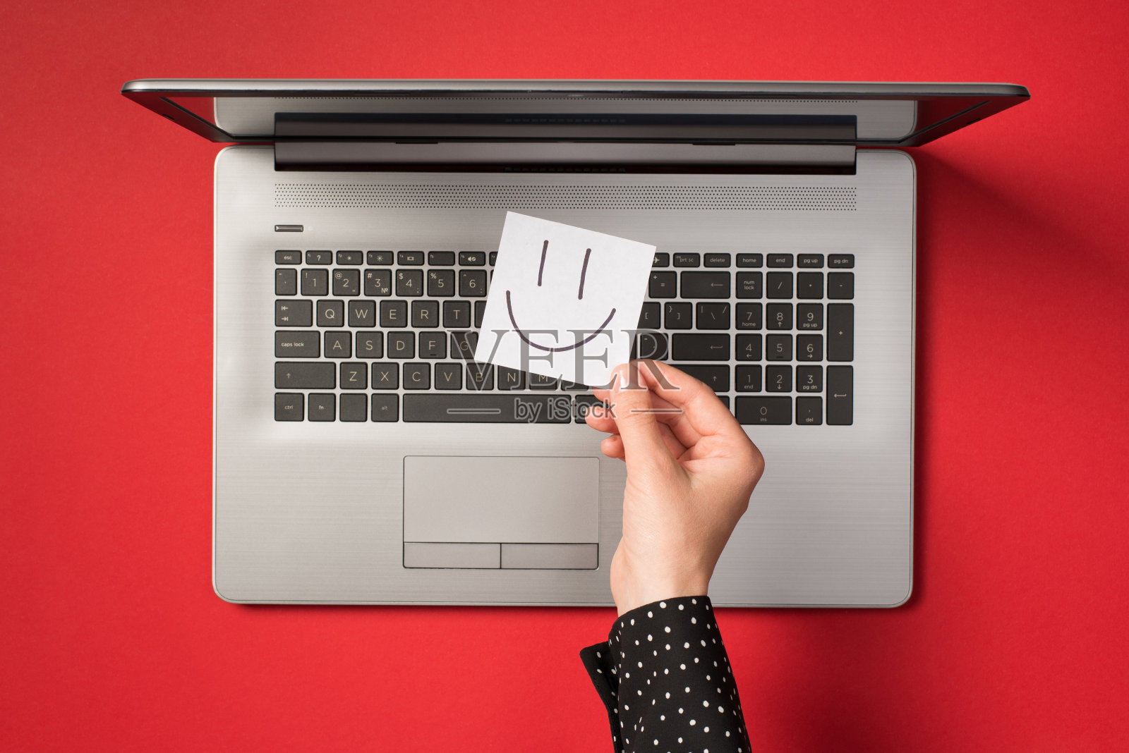第一个人俯视图的照片，手握白色贴纸纸条与画的微笑的脸在开放的灰色笔记本电脑孤立的红色背景照片摄影图片